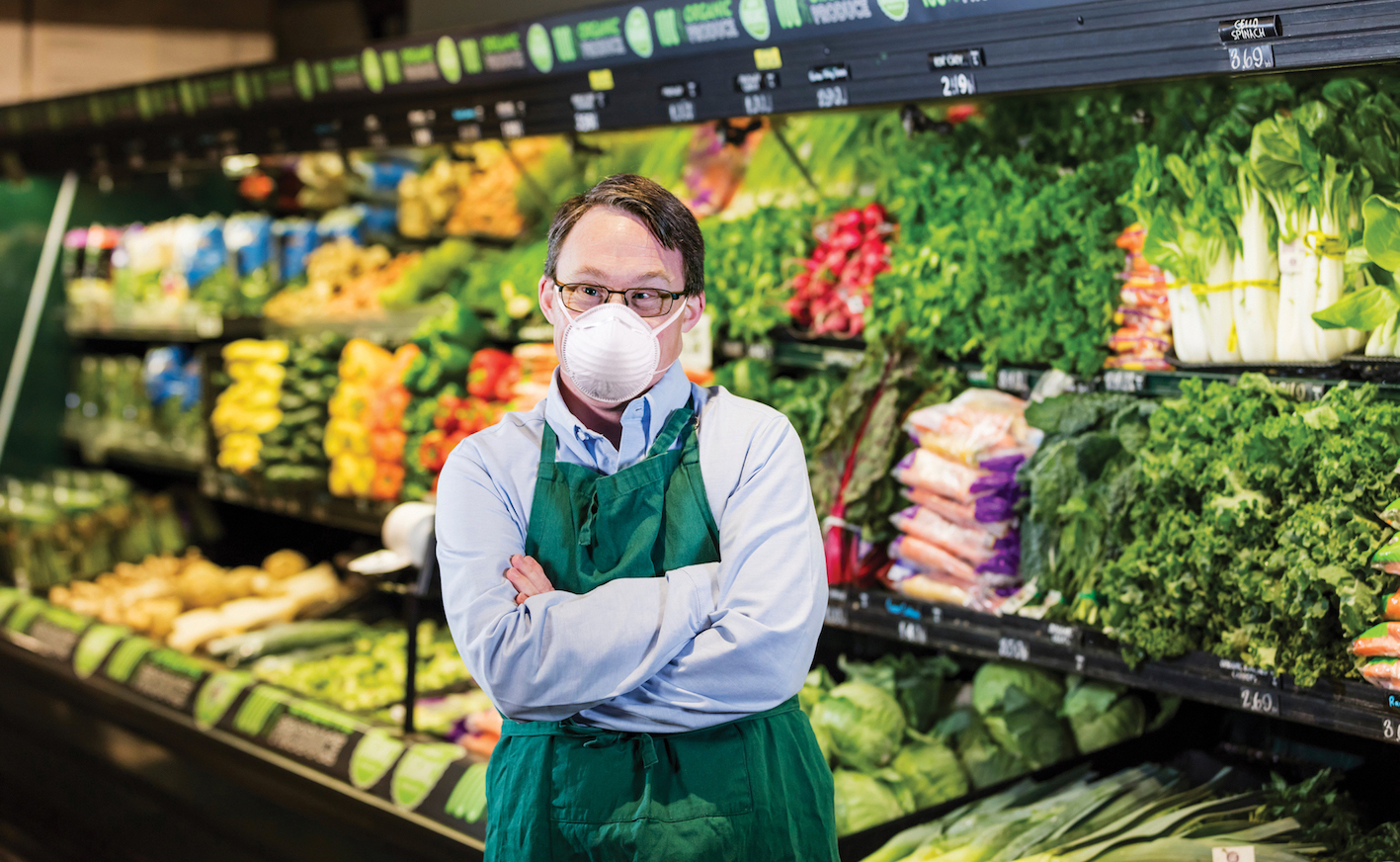 A male grocery store worker wearing a mask and standing in front of vegetables.