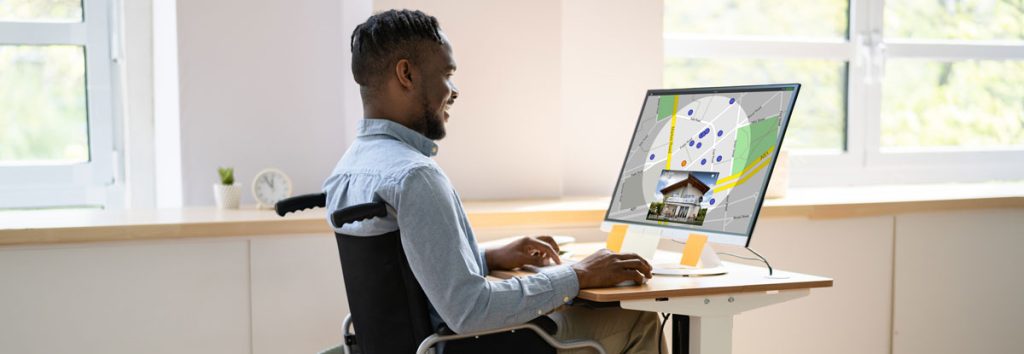 A professional black man in a manual wheelchair sits at an adapted desk working on a computer. The monitor shows a map with several points marked on it.
