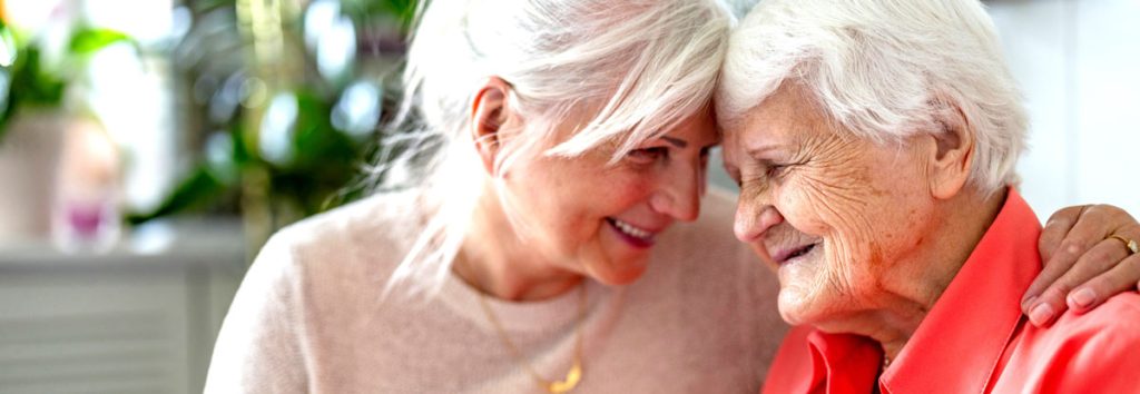 Two older women sit smiling with their heads together.