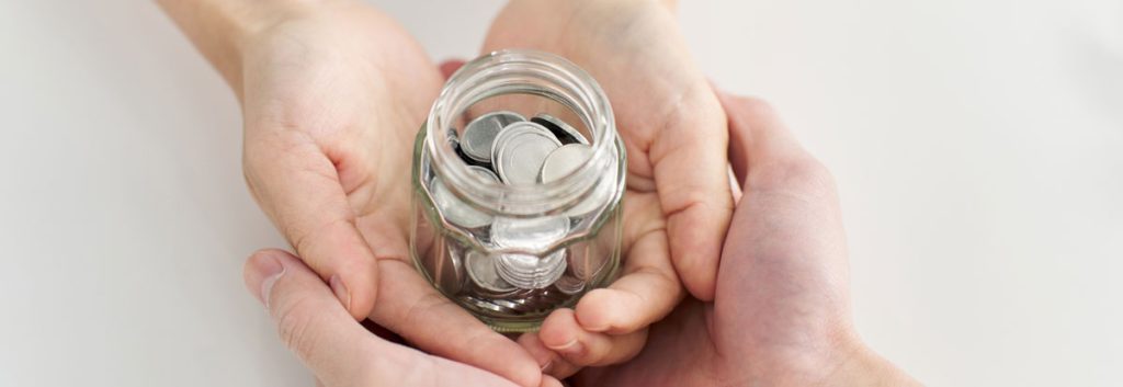 Two sets of hands, one atop the other, cradle a small glass jar filled with change