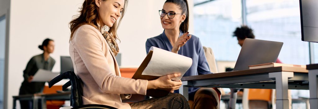 A professional woman in a manual wheelchair sits at a table in a busy office discussing paperwork with another professionally dressed woman who is working on a laptop.