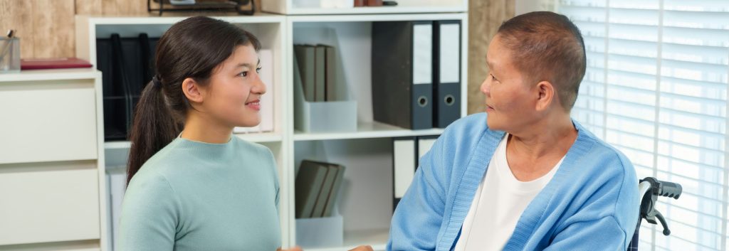 A young woman sits facing an older woman who is in a wheelchair, and has very thin, short hair, possibly from medical treatment.